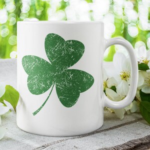 May include: White ceramic mug featuring a distressed green shamrock design. The shamrock is a three-leaf clover, a symbol of luck and Irish heritage. The mug has a curved handle and is set against a blurred green background.