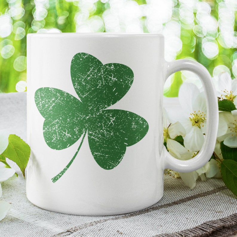 May include: White ceramic mug featuring a distressed green shamrock design. The shamrock is a three-leaf clover, a symbol of luck and Irish heritage. The mug has a curved handle and is set against a blurred green background.