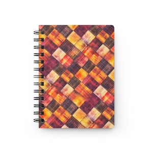 Puede incluir: Cuaderno con espiral con un patrón geométrico colorido. La cubierta presenta un diseño repetitivo de cuadrados en tonos de rojo, naranja, amarillo y marrón, creando una apariencia texturizada similar a un tartán. La encuadernación en espiral del cuaderno es negra.