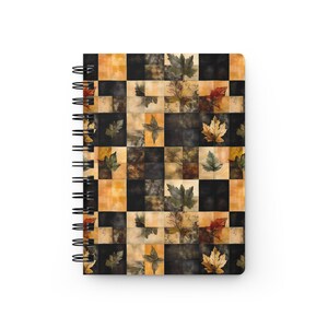 May include: Spiral-bound notebook featuring a fall-themed design. The cover showcases a patchwork pattern of squares in black, tan, and orange hues, adorned with leaf illustrations in shades of green, brown, and red. The notebook's spiral binding is black.