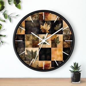 Puede incluir: Reloj de pared redondo con marco negro. La esfera presenta un patrón de cuadros con hojas de otoño en tonos marrón, verde y dorado. Las manecillas y los marcadores blancos indican la hora. Un elemento decorativo.
