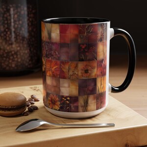May include: A ceramic coffee mug with a mosaic design of flowers and foliage in shades of orange, brown, and purple. The mug has a black handle and rim, and a white interior. The mug is on a wooden surface.