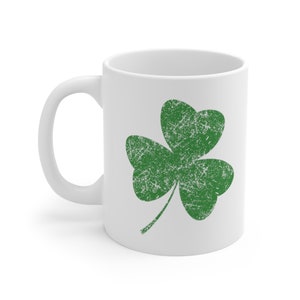 May include: White ceramic mug featuring a distressed green shamrock design. The shamrock is a three-leaf clover, a symbol of luck and St. Patrick's Day. The mug has a curved handle for easy gripping.