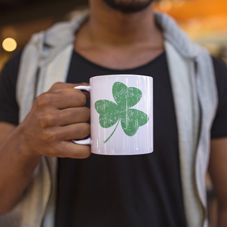 A young man models an 11-ounce white ceramic coffee mug featuring Sweet Holdings distressed, green shamrock, design, printed on both the front and back sides of the mug.