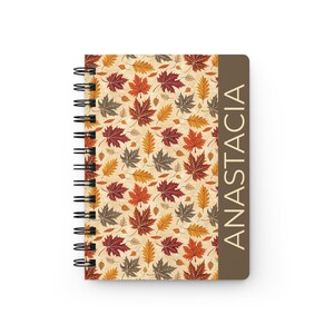 Puede incluir: Cuaderno de espiral con un patrón de hojas de otoño en tonos rojos, naranjas y marrones sobre un fondo crema. El lomo es negro y el lado derecho tiene el nombre "ANASTACIA" impreso verticalmente en un rectángulo marrón.