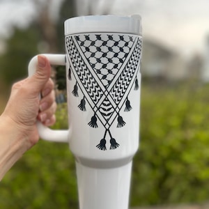 May include: White travel mug with a black and white patterned design. The pattern is a traditional Palestinian keffiyeh.