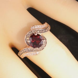 May include: A rose gold ring featuring a large, round, deep red gemstone at the center, surrounded by smaller, clear stones. The band has a swirling design with additional small stones.
