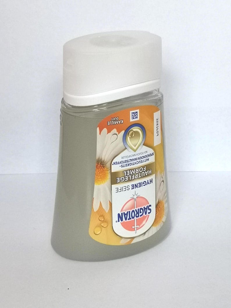 Puede incluir: Una botella transparente en forma de pera de jab&oacute;n de higiene Sagrotan con tapa blanca. La etiqueta presenta el nombre de la marca, detalles del producto y un dise&ntilde;o de flor de manzanilla. La botella contiene un l&iacute;quido gris claro.