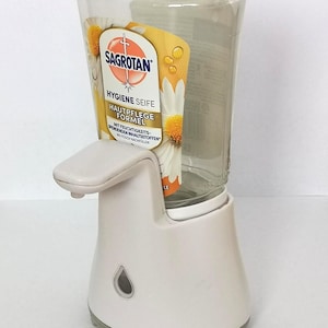 Puede incluir: Dispensador de jab&oacute;n autom&aacute;tico blanco y transparente con boquilla curva y base. El dispensador tiene un dep&oacute;sito de jab&oacute;n de pl&aacute;stico transparente con una etiqueta que dice "SAGROTAN HYGIENE SEIFE".
