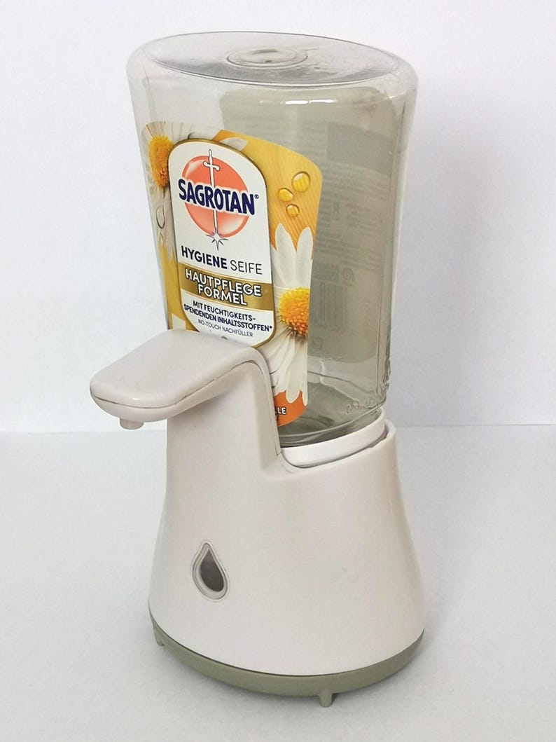 Puede incluir: Dispensador de jab&oacute;n autom&aacute;tico blanco y transparente con boquilla curva y base. El dispensador tiene un dep&oacute;sito de jab&oacute;n de pl&aacute;stico transparente con una etiqueta que dice "SAGROTAN HYGIENE SEIFE".