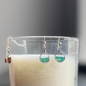 May include: Silver-tone earrings with a half-circle design, featuring a turquoise and green speckled inlay. The earrings are suspended from a silver hook. Another pair of earrings with a teardrop shape and brown inlay is also visible.