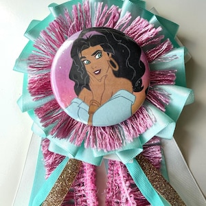 May include: A circular pinback button featuring an illustration of a woman with long dark hair, gold hoop earrings, and a light blue off-the-shoulder top. The button is centered on a layered rosette made of light blue ribbon and pink fringe.