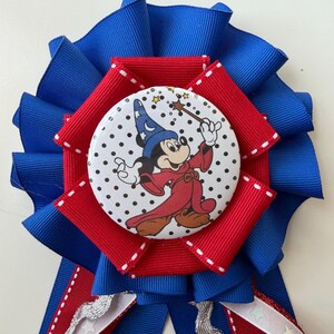 May include: A circular award ribbon featuring a central button with a graphic of a cartoon mouse in a blue wizard hat and red robe holding a wand. The button is set against pleated blue and red ribbons with white stitching and dangling streamers.
