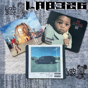 May include: Three different square coasters featuring album covers. The first coaster features a cartoon image of a man with a rocket ship in the background. The second coaster features a young boy with a black and white design on his face. The third coaster features a black minivan against a blue sky. The text on the coasters reads "Lil Wayne Tha Carter III" and "gooo KOO MANO CITY ACT ADVISORY BY HOODZ GAMMA".