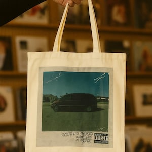 May include: Cream-colored tote bag featuring a graphic of a black van on a road under a blue sky. The bag has long handles and text at the bottom. The image has a vintage aesthetic. The bag is held up by a person.