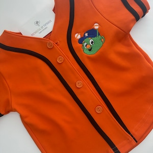 May include: Orange baseball jersey with black stripes and a green alien mascot with a baseball cap and bat. The jersey has a button-up front and the text "Pocketful of Threads Custom Embroidery" on the left side.