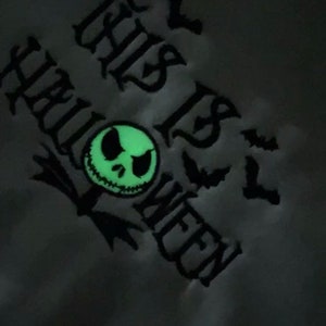 May include: A black and white graphic design of a Jack Skellington face with the text "This is Halloween" in a gothic font. The design is on a gray background and glows in the dark.