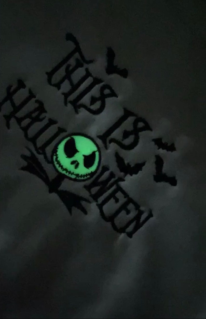 May include: A black and white graphic design of a Jack Skellington face with the text "This is Halloween" in a gothic font. The design is on a gray background and glows in the dark.