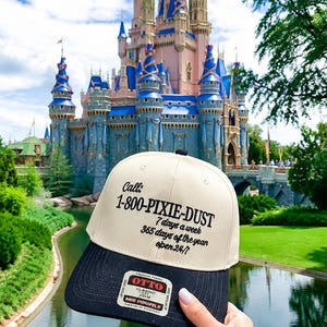 Puede incluir: Una gorra de béisbol bicolor con panel frontal color crema y visera negra. El frente presenta texto negro que dice: Call: 1-800-PIXIE-DUST, 7 days a week, 365 days of the year, open 24/7. Una pegatina blanca con texto negro está en la visera.