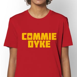 May include: A red t-shirt with the text "COMMIE DYKE" in yellow block letters. The shirt has a crew neck and short sleeves. The image also includes a "GOTS" and "Vegan" logo.