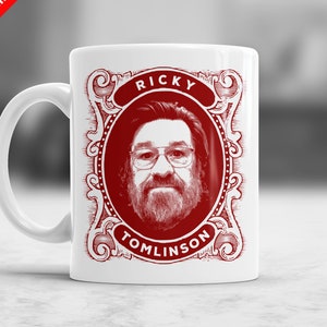 May include: White ceramic mug with a red portrait of a man with glasses and a beard, framed by an ornate design. The name "RICKY TOMLINSON" is printed above and below the portrait. The mug has a curved handle.