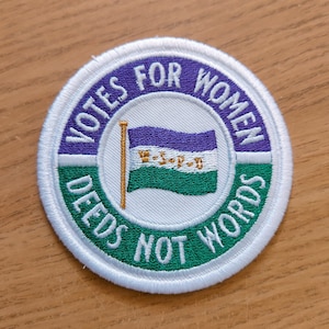 Może przedstawiać: Okrągła biała naszywka z zielonym i fioletowym obramowaniem. Naszywka przedstawia białą flagę z fioletowym i zielonym wzorem oraz tekstem "Votes for Women Deeds Not Words".