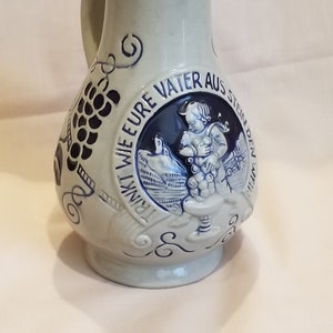May include: A light blue ceramic pitcher with a handle and a spout. The pitcher features a dark blue embossed design of grapes and a circular emblem with a figure and text. The text reads "TRINKT WIE EURE VATER AUS STEIN DEN WEIN."