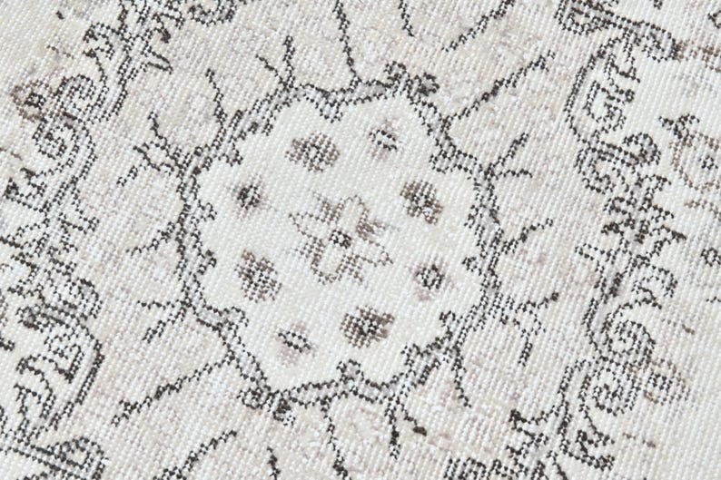 Peut inclure: Gros plan sur un tapis blanc avec un motif g&eacute;om&eacute;trique noir et gris. Le motif pr&eacute;sente un m&eacute;daillon central avec des motifs floraux r&eacute;p&eacute;t&eacute;s.