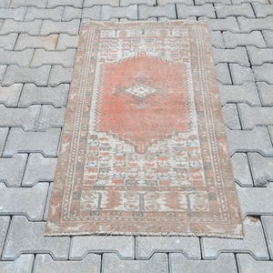 Puede incluir: Alfombra rectangular de estilo vintage con un medallón central en tonos melocotón y crema. La alfombra presenta intrincados diseños de borde en tonos apagados, con un aspecto desgastado. Se encuentra sobre una superficie gris texturizada.
