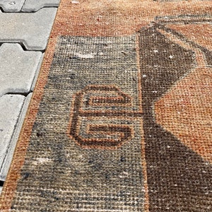 Pu&ograve; includere: Un primo piano di un tappeto vintage con un motivo geometrico in tonalit&agrave; di marrone, arancione e grigio. Il tappeto ha un aspetto usurato e sbiadito, che gli conferisce un aspetto rustico e antico.