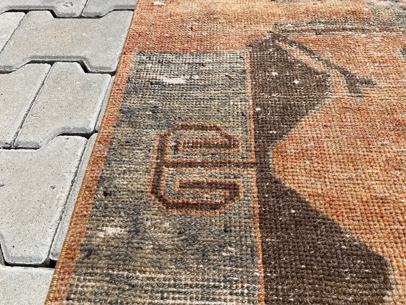 Pu&ograve; includere: Un primo piano di un tappeto vintage con un motivo geometrico in tonalit&agrave; di marrone, arancione e grigio. Il tappeto ha un aspetto usurato e sbiadito, che gli conferisce un aspetto rustico e antico.