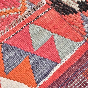 May include: Close-up of a colorful woven rug with a geometric pattern of triangles in shades of red, orange, gray, and black. The rug has a textured surface and a fringe border.