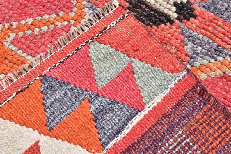 May include: Close-up of a colorful woven rug with a geometric pattern of triangles in shades of red, orange, gray, and black. The rug has a textured surface and a fringe border.