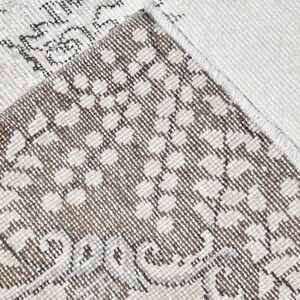 Peut inclure: Gros plan sur un tapis beige et brun &agrave; motifs avec une bordure blanche. Le tapis a une texture tiss&eacute;e et un motif floral subtil.