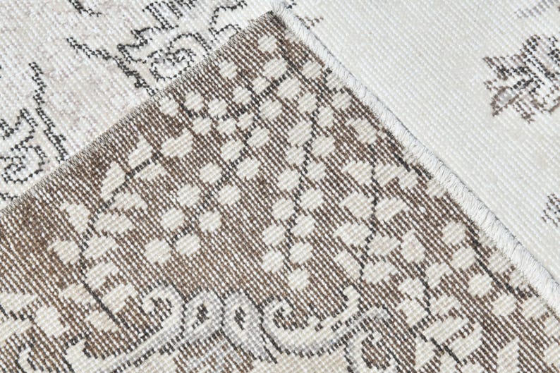 Peut inclure: Gros plan sur un tapis beige et brun &agrave; motifs avec une bordure blanche. Le tapis a une texture tiss&eacute;e et un motif floral subtil.