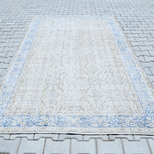 May include: A rectangular beige rug with a floral pattern and a blue border, laid on a grey brick surface. The rug's design features a central field with a repeating floral motif.