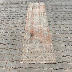 May include: A long, rectangular rug with a faded, distressed look. The rug has a light beige background with a subtle pattern of faded pink and brown. The rug is lying on a grey concrete surface.