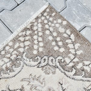Peut inclure: Gros plan sur un tapis beige et brun &agrave; motifs avec une bordure en franges. Le tapis est pos&eacute; sur une surface en b&eacute;ton gris.