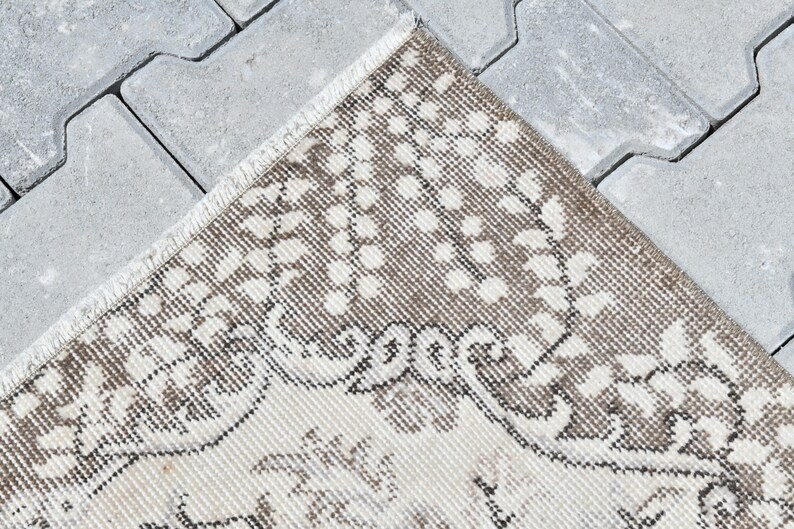 Peut inclure: Gros plan sur un tapis beige et brun &agrave; motifs avec une bordure en franges. Le tapis est pos&eacute; sur une surface en b&eacute;ton gris.