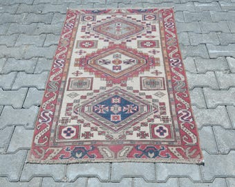 May include: Rectangular, hand-woven rug with a geometric pattern. The rug features a cream-colored background with red, blue, and brown geometric shapes. The border is red with a repeating pattern. The rug is placed on a gray stone surface.