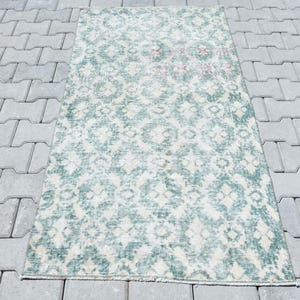 Può includere: Un tappeto in stile vintage con sfondo bianco e un motivo geometrico verde. Il tappeto è realizzato in un materiale morbido e tessuto e ha un aspetto vissuto.