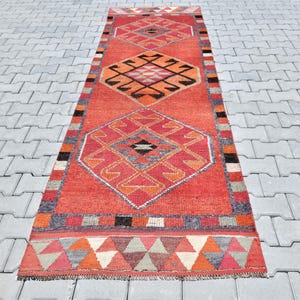May include: A long, red and orange runner rug with a geometric pattern. The rug has a central diamond shape with smaller diamonds within it. The rug has a fringe border.