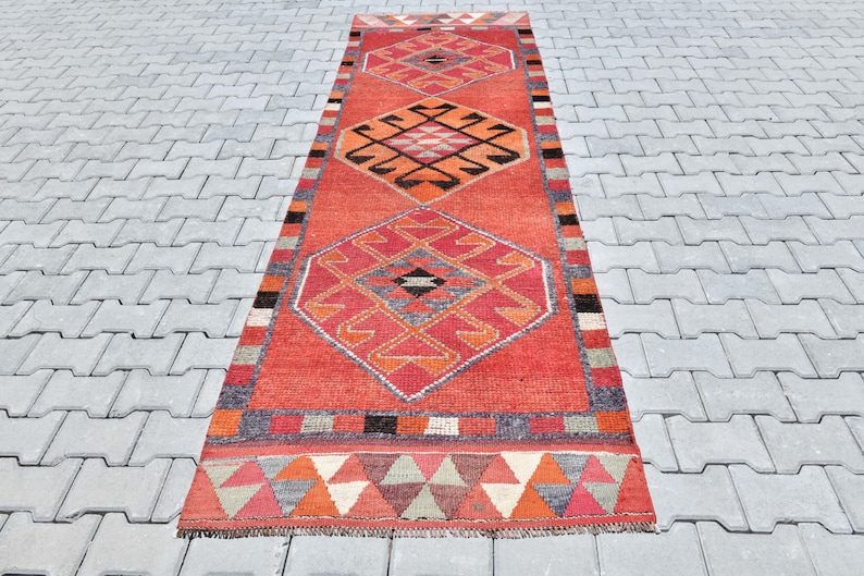 May include: A long, red and orange runner rug with a geometric pattern. The rug has a central diamond shape with smaller diamonds within it. The rug has a fringe border.