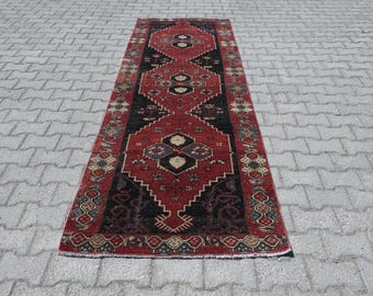 May include: A rectangular, vintage-style rug with a central design in red and black. The rug features geometric patterns and a border with floral motifs. The rug is placed on a gray, textured surface.