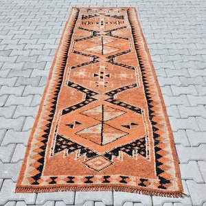 Könnte beinhalten: Ein langer, schmaler Teppich mit einem geometrischen Muster in Orange-, Schwarz- und Weißtönen. Der Teppich hat an beiden Enden Fransen.