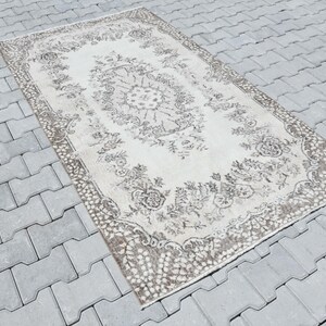 Peut inclure: Un tapis vintage avec un motif floral dans des tons beige et marron. Le tapis a un aspect vieilli et est pos&eacute; sur une surface en b&eacute;ton gris.