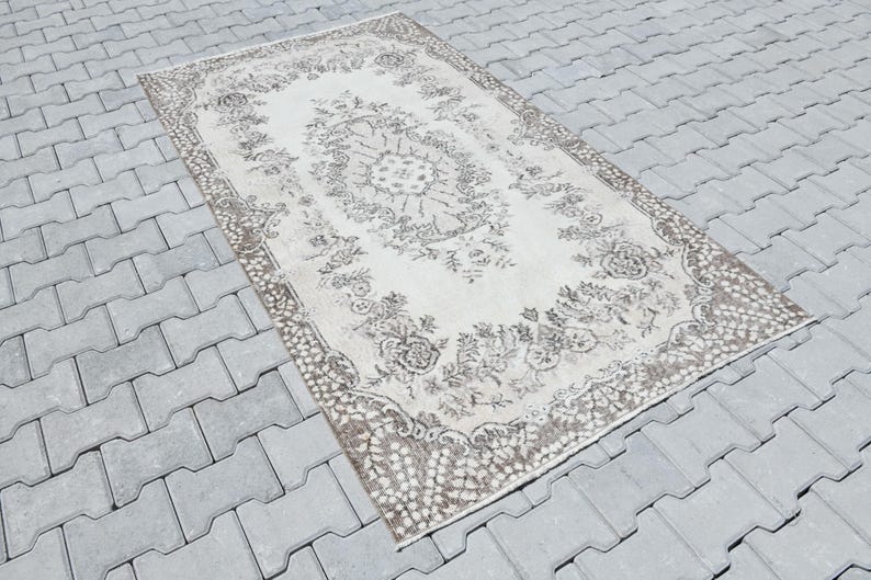 Peut inclure: Un tapis vintage avec un motif floral dans des tons beige et marron. Le tapis a un aspect vieilli et est pos&eacute; sur une surface en b&eacute;ton gris.