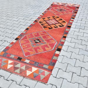 May include: A long, rectangular red rug with a geometric pattern in shades of orange, pink, and black. The rug has a fringe border and is laid out on a gray concrete surface.
