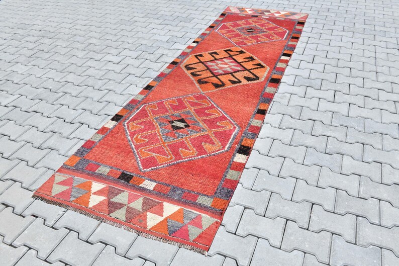 May include: A long, rectangular red rug with a geometric pattern in shades of orange, pink, and black. The rug has a fringe border and is laid out on a gray concrete surface.