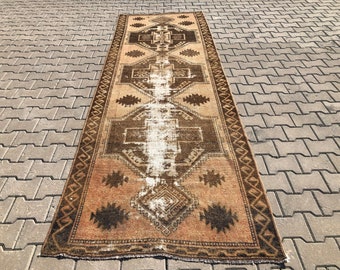 May include: A vintage runner rug with a brown and beige color scheme. The rug has a geometric pattern with a central diamond shape and a border of repeating motifs.